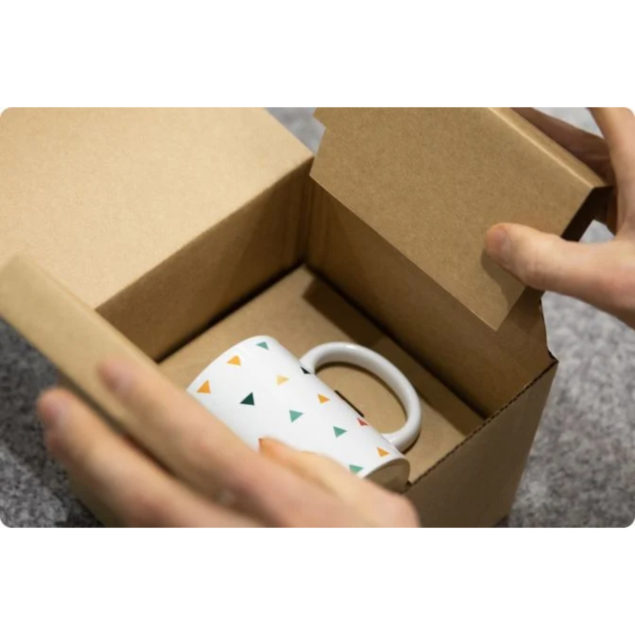 White mug with colorful triangle pattern inside an open cardboard box, held by a hand.